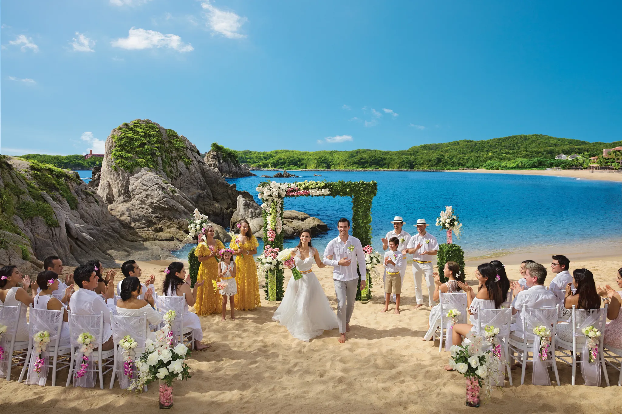 Beach wedding ceremony by rocky formations with floral arch at Dreams Huatulco Resort & Spa