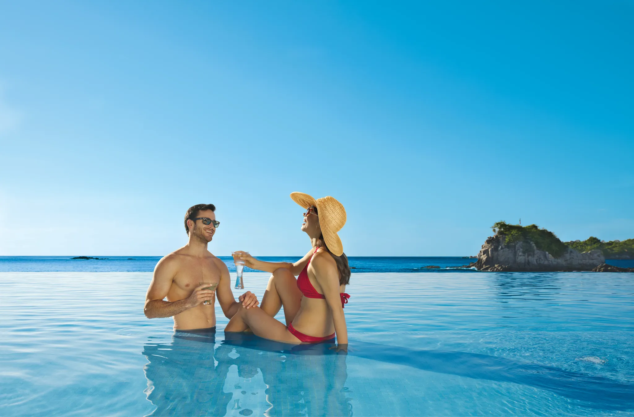 Couple relaxing in the infinity pool with cocktails at Dreams Huatulco Resort & Spa overlooking the ocean.